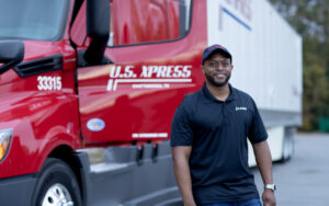 An African-American U.S. Xpress driver wearing a black shirt and hat smiles while a U.S. Xpress truck sits in the background.