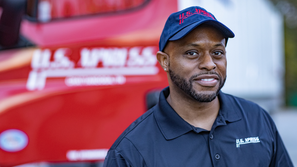 U.S. Xpress Driver wearing blue shirt and hat in front of red U.S. Xpress truck.